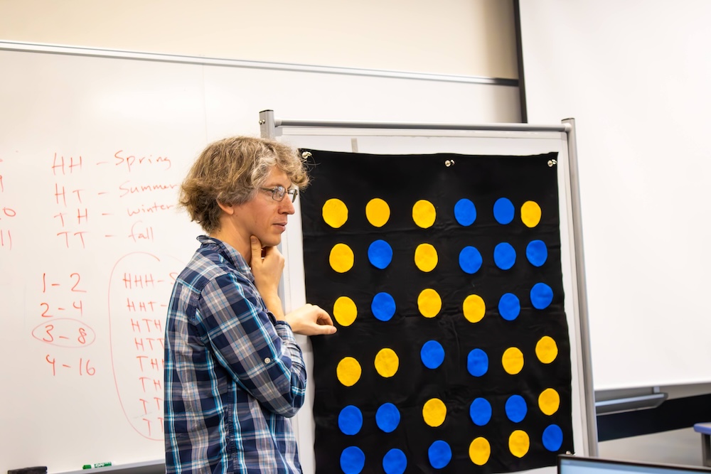 Dr. Johnson stands beside a large black board arranged with blue and yellow circular markers, with handwritten notes on a whiteboard behind them.