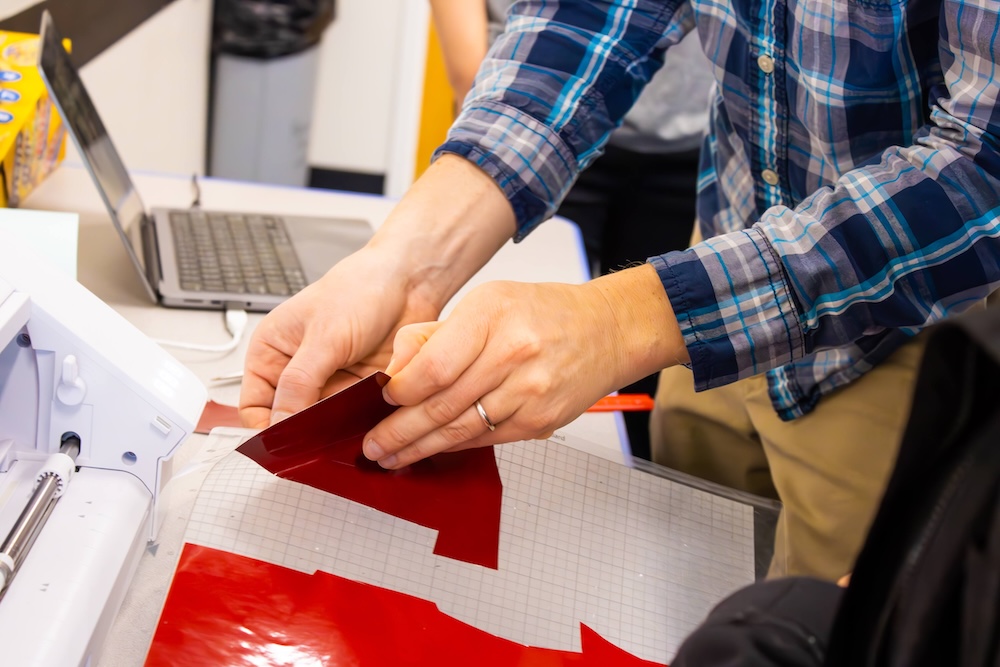 Hands peeling a red vinyl sheet on a cutting mat beside a machine, with a laptop on the desk in the background.