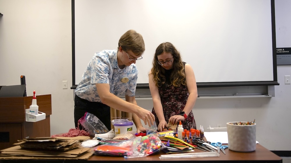Two people work together at a table covered with craft supplies, with a projector screen behind them in a classroom.