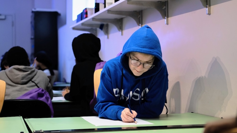 Student in a blue hoodie and glasses writes on paper at a classroom desk while other students sit in the background.