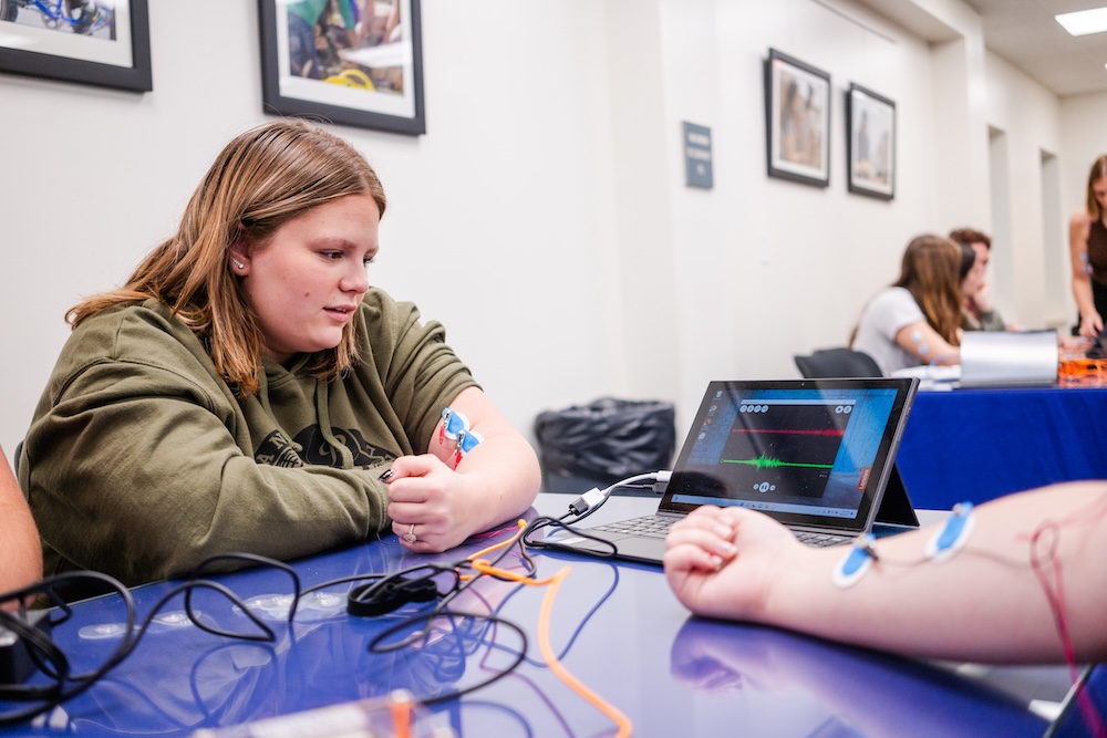 Student watches a laptop display of sensor readings during a lab activity, with electrodes attached to a person’s arm on the table.