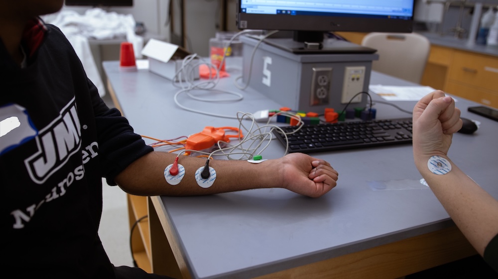 Two people at a lab table with electrode pads attached to their forearms and wires connected to monitoring equipment.