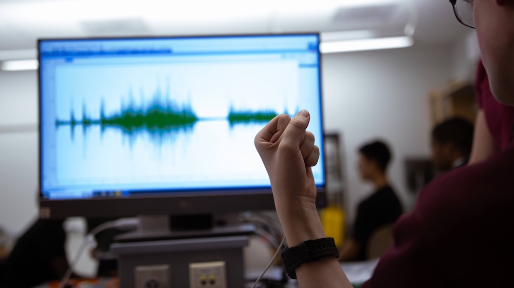 Person gestures with a fist while looking at a computer monitor displaying an audio waveform, with a blurred classroom setting in the background.