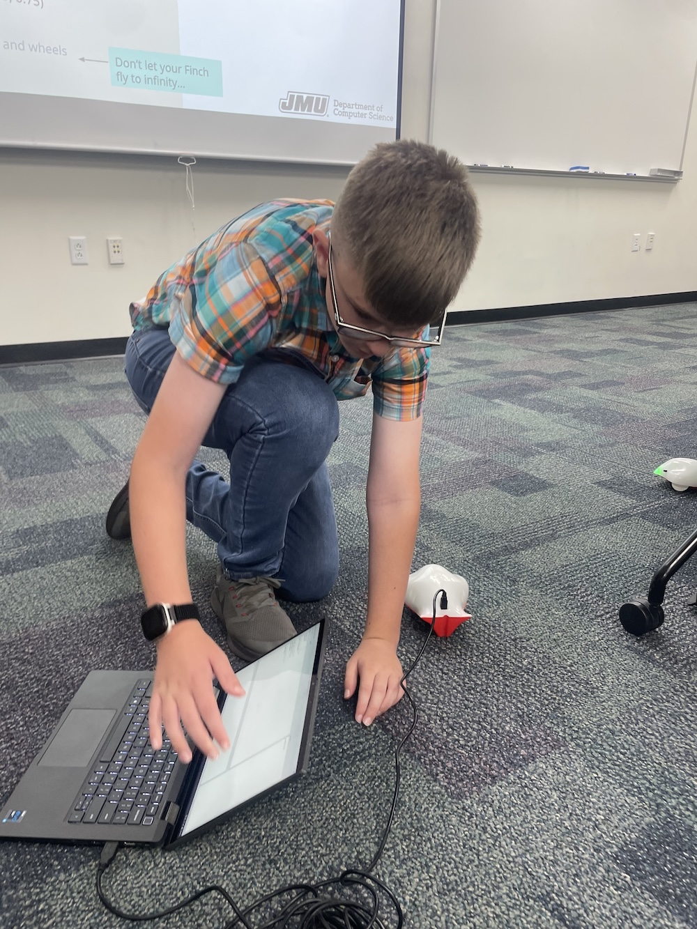 Studen kneels on a classroom floor adjusting a cable connecting a laptop to a finch robot.