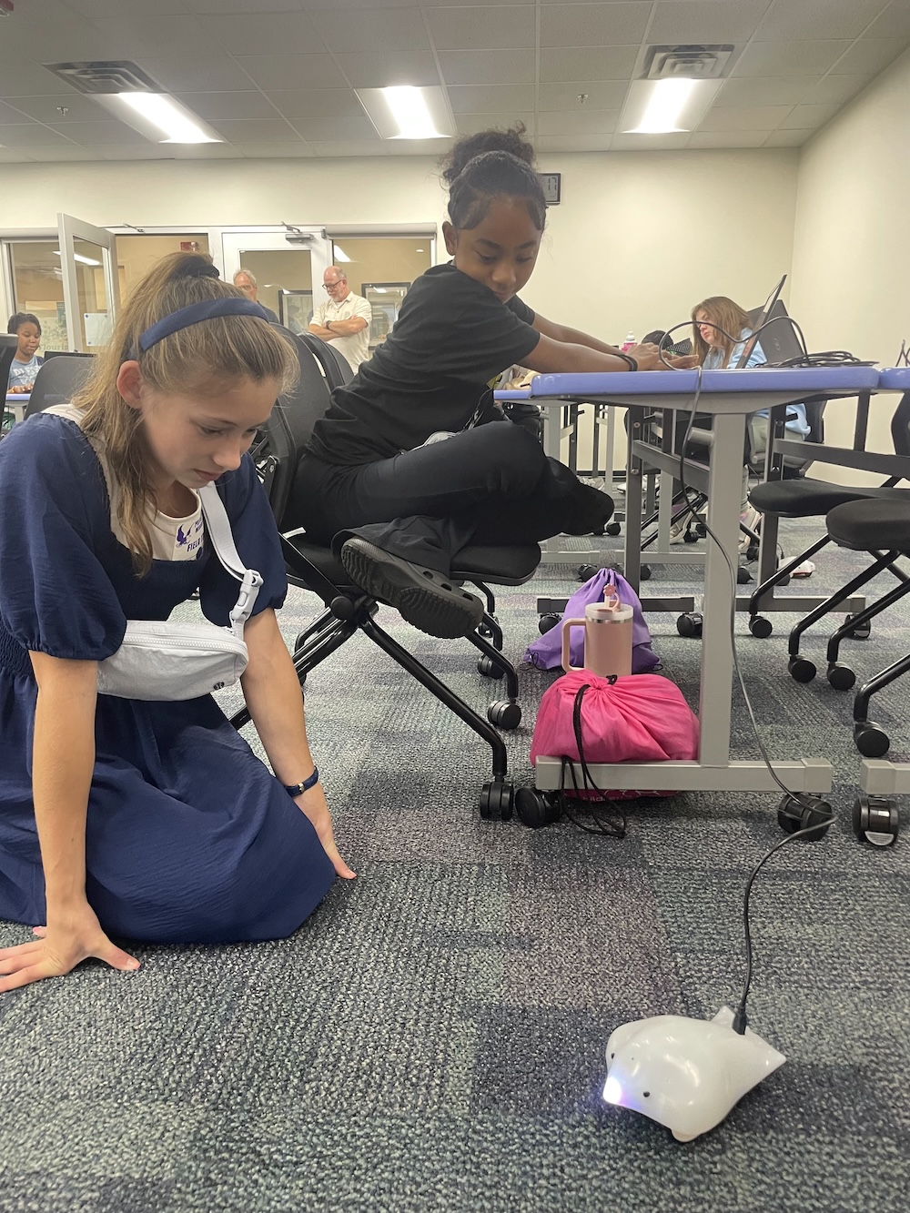 Two students observe a finch robot with a glowing nose as it moves across the carpet.