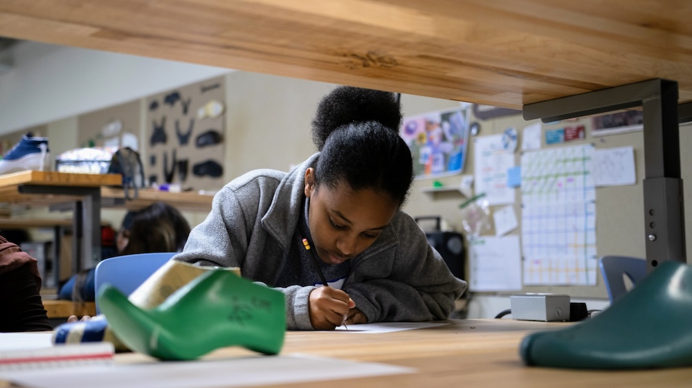 Student leaning over a desk and writing on paper in a classroom workshop with shoes and posters in the background.