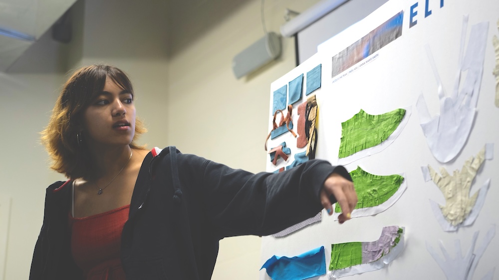 Student points to a classroom presentation board covered with colorful paper cutouts and notes.