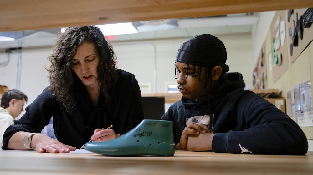 Instructor helps a student at a workbench, with a green shoe last in the foreground.