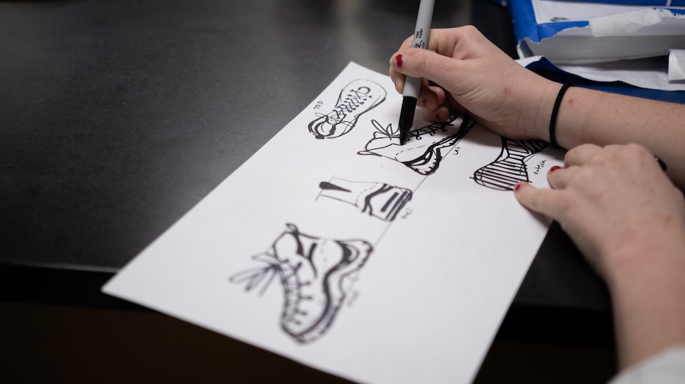 Hands sketching sneaker designs with a black marker on a sheet of paper at a desk.