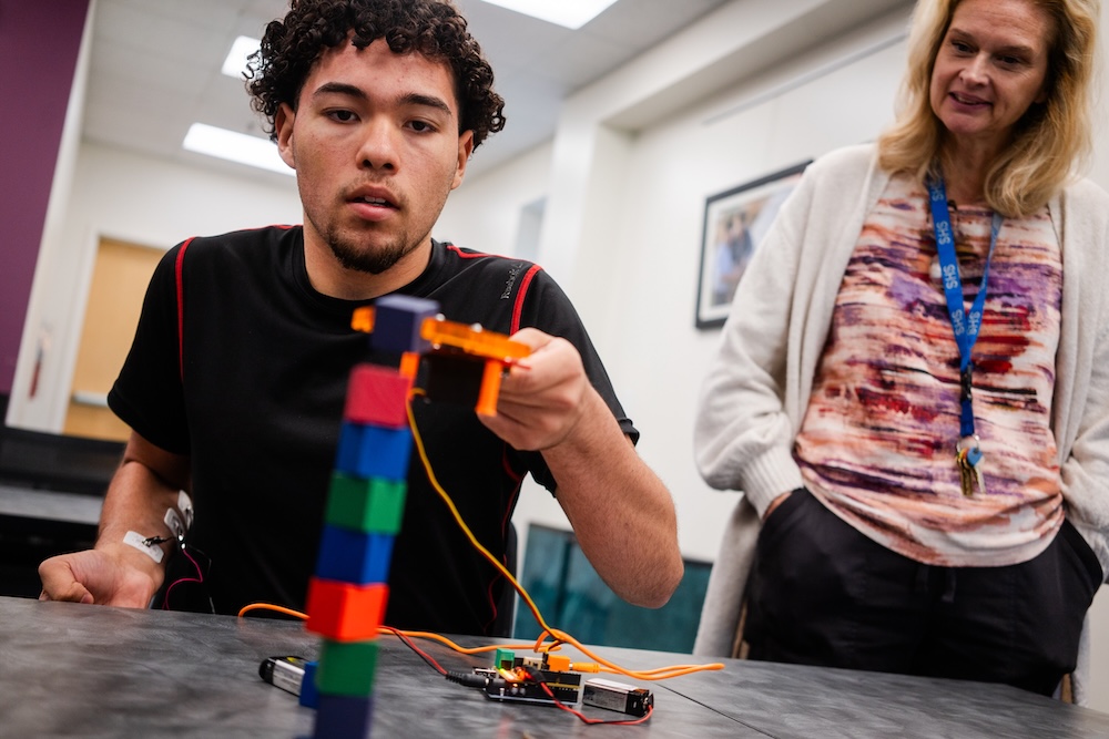 Student adjusts a small robotic device with wires and a circuit board on a table while an instructor watches nearby.