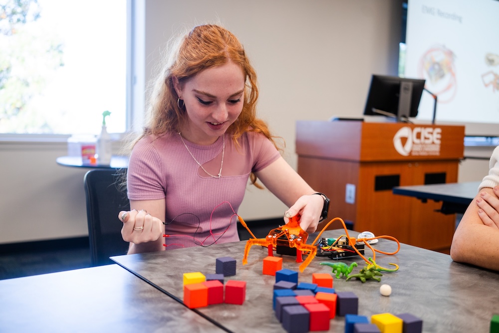 Student in a classroom tests a small orange walking robot on a table scattered with colorful blocks and wires.