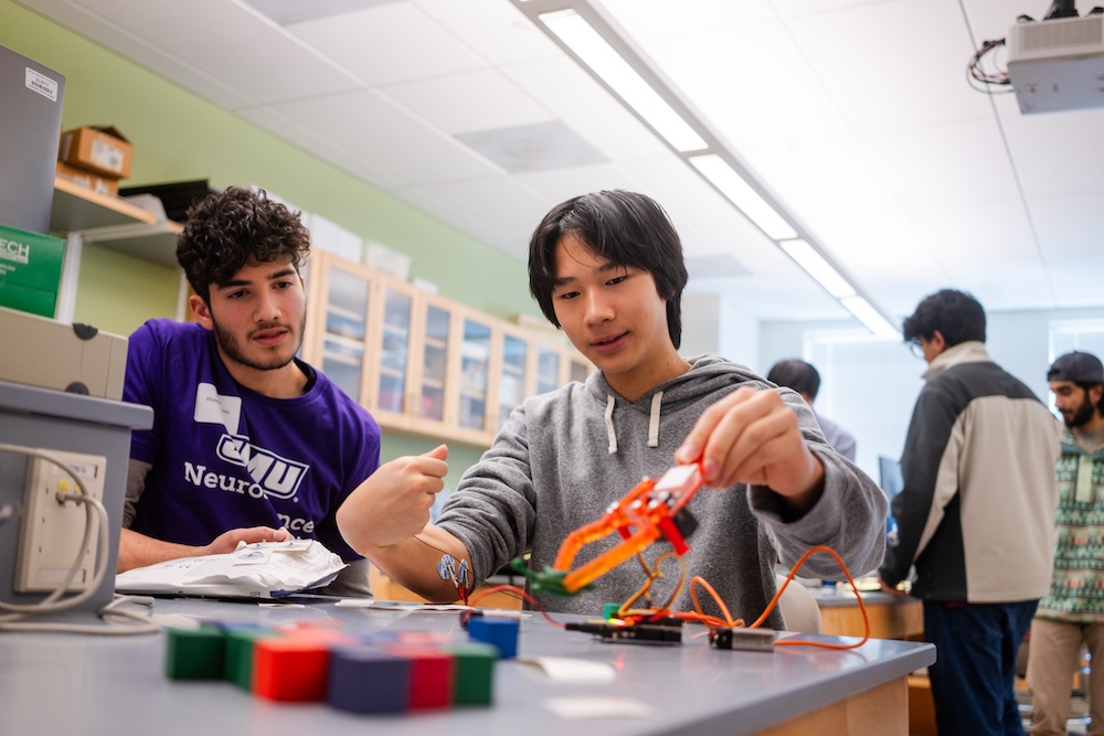 Two students work together at a classroom lab table, assembling an electronics project with wires and a small robotic arm while other students are in the background.