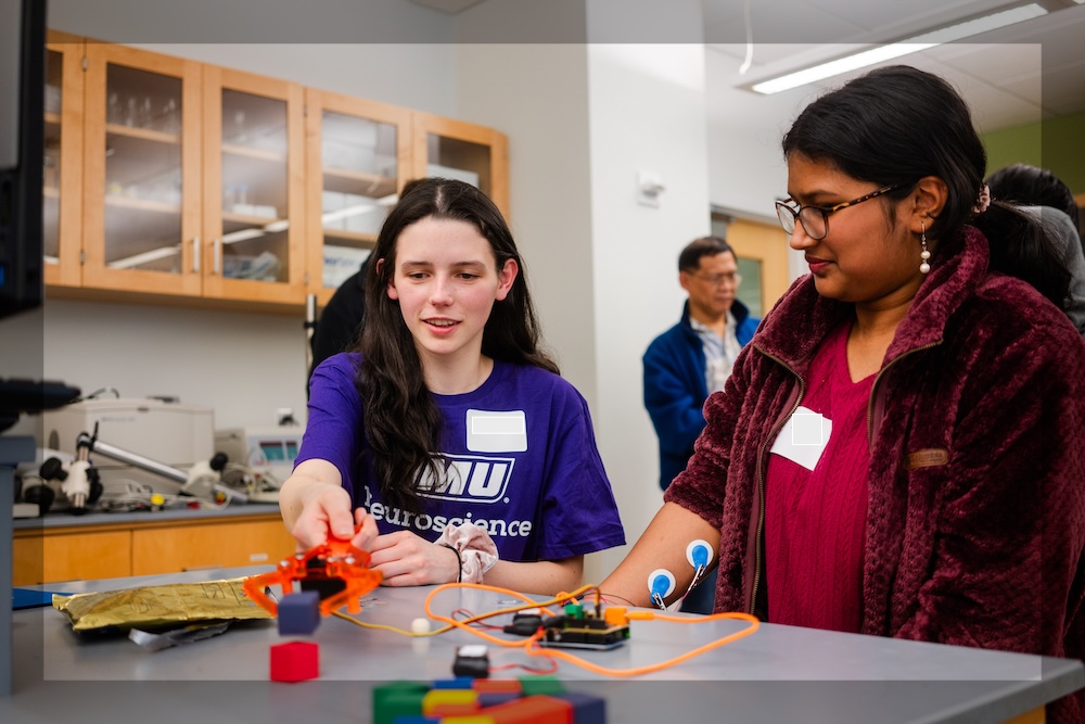 Two students in a science lab work together at a table with wires, sensors, and a small device during a hands-on experiment.