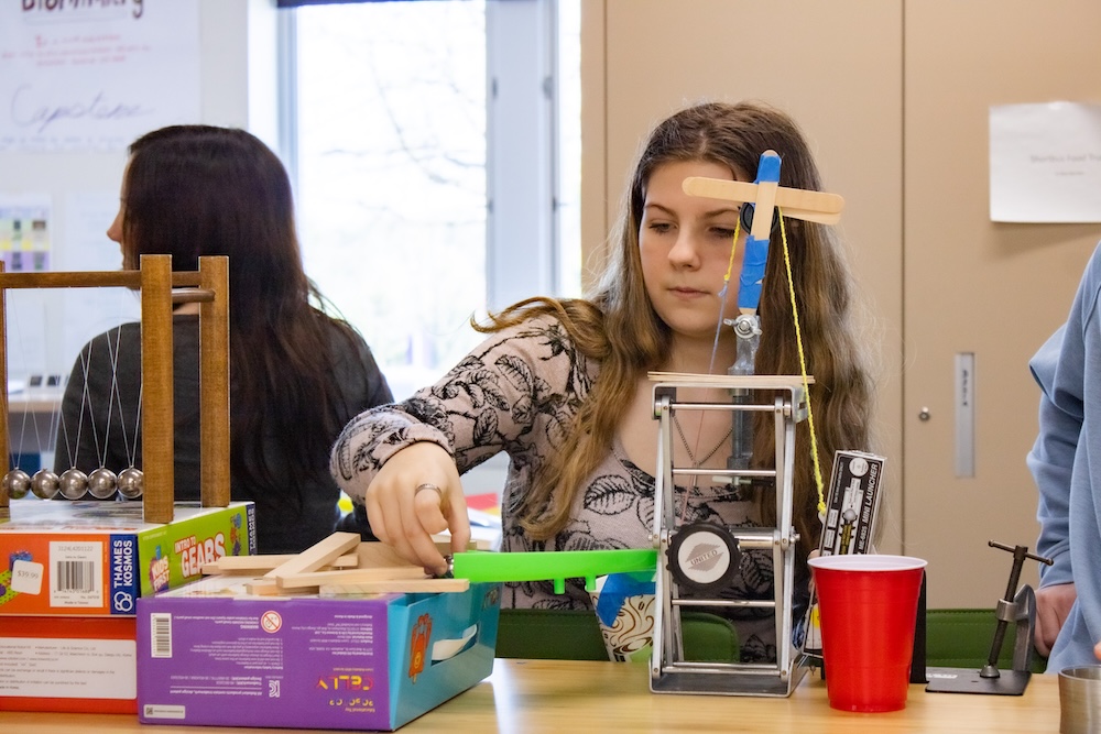 Student works at a building a Rube Goldberg Machine with a small pulley and lever setup made from craft sticks and string. 