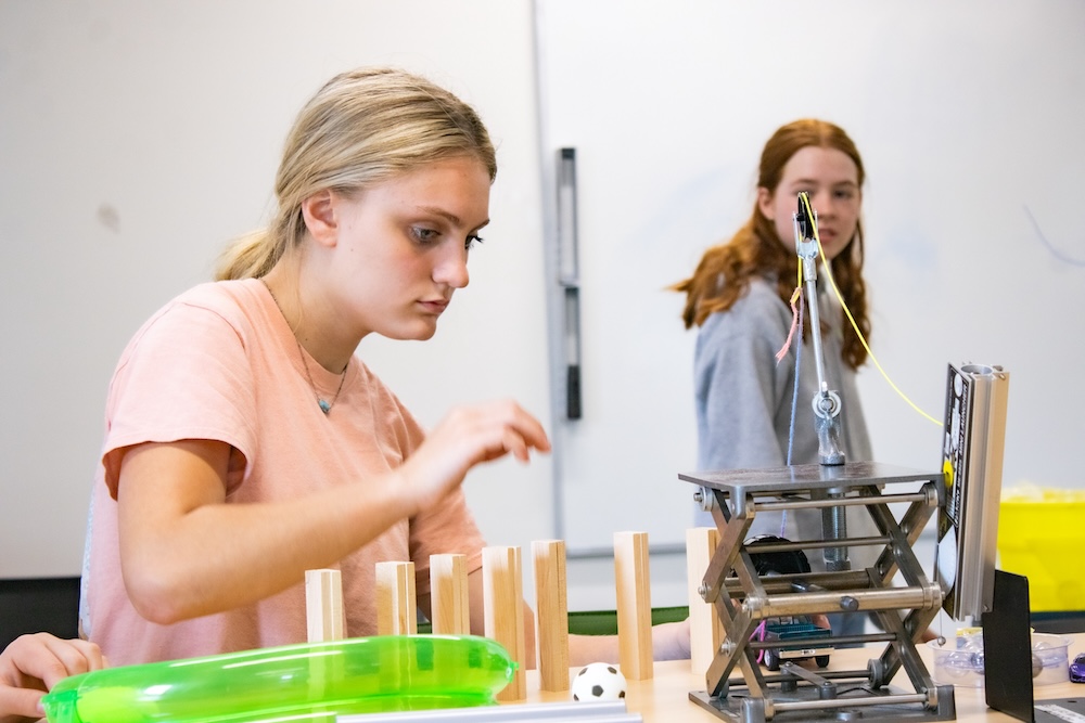 Student concentrates while assembling a small engineering setup with wooden blocks and a metal scissor-lift mechanism on a classroom table.