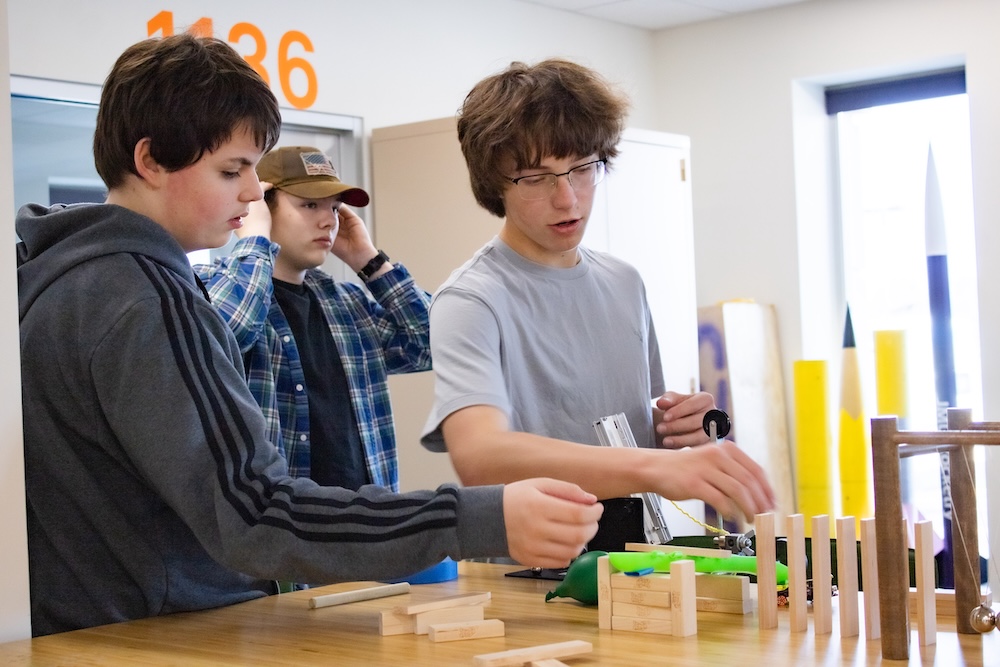 Three students work together at a classroom workbench, assembling a small wooden model with measuring tools and parts spread out on the table.