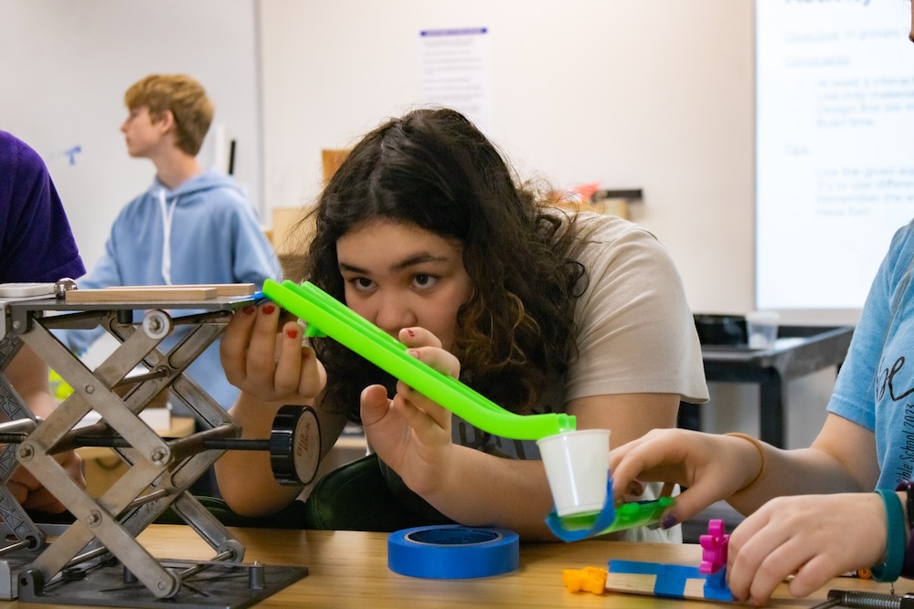 Student adjusts a bright green plastic ramp over a table as another student steadies the end with a small cup, in a classroom engineering activity.