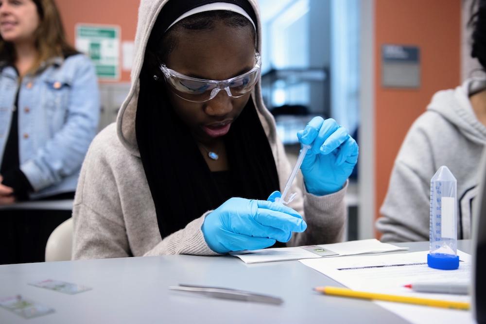 Student wearing safety goggles and blue gloves uses a pipette to transfer liquid during a science lab activity at a classroom table.