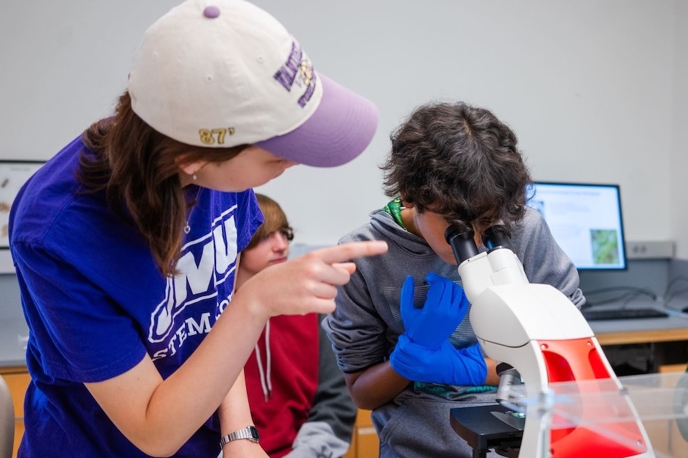 Student points while another student wearing blue gloves looks through a microscope in a classroom lab.