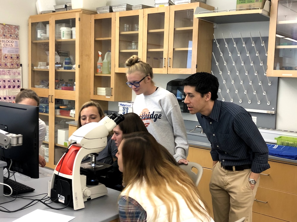 Students and an instructor gather around a microscope and computer screen in a science lab classroom.
