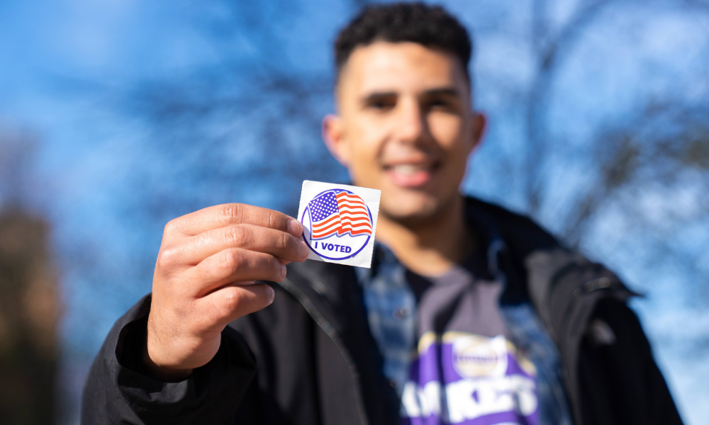 <span class="submesa"><span class="slidetitle">2022 Election</span></span> Civic fellow showing the camera his "I voted" sticker
