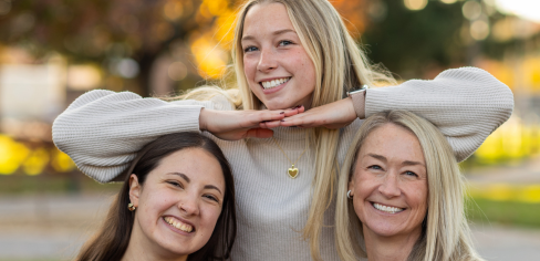 Alli resting her arms on Margaret and Paige's heads while they sit on a bench in the Fall on campus