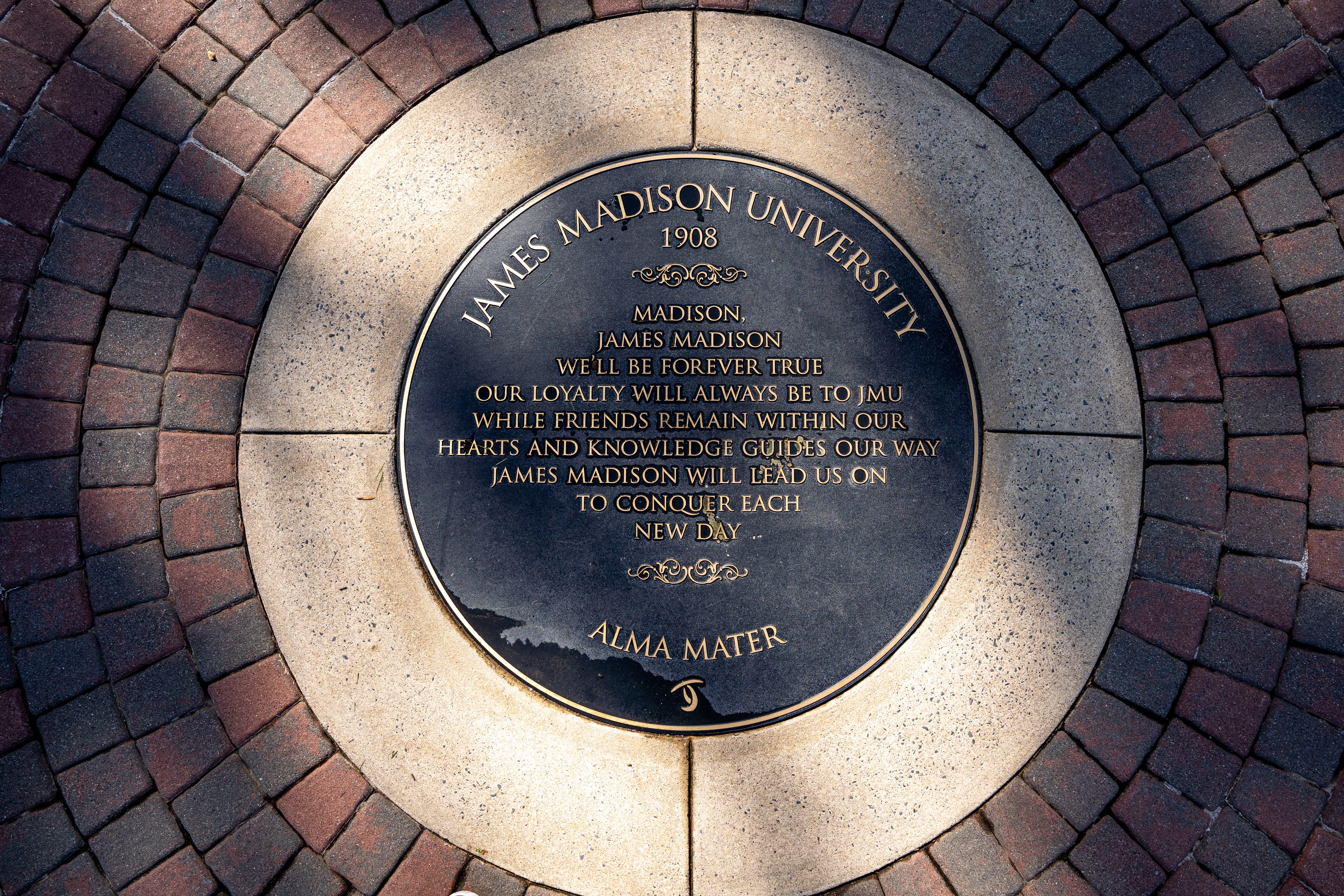 Circular James Madison University alma mater plaque embedded in a brick plaza, with gold lettering and the year 1908.