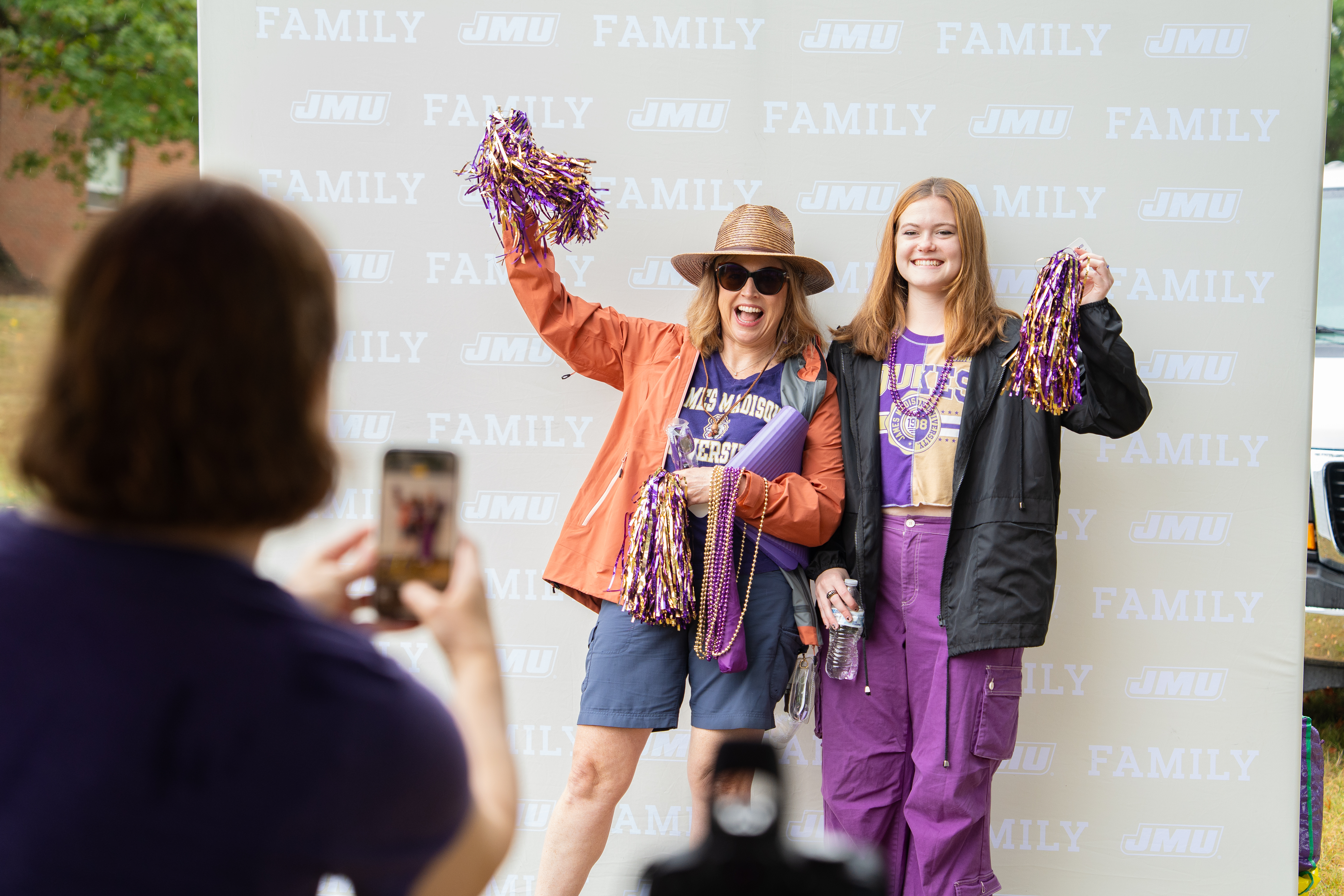 Two people pose smiling with purple-and-gold pom-poms in front of a JMU Family step-and-repeat backdrop while someone in the foreground takes their photo.